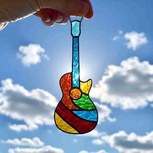 May include: A colorful stained glass guitar pendant with a blue neck and headstock, and a body of red, orange, yellow, green, and blue glass. The pendant is held up against a bright blue sky with fluffy white clouds.