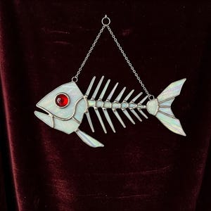 May include: A stained glass fish skeleton suncatcher with a red eye and iridescent fins. The decorative piece hangs from a silver chain. The fish skeleton is a unique home decor item.