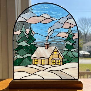 May include: A stained glass window depicting a cozy cabin in a snowy winter scene. The cabin has a yellow exterior, a white roof, and smoke coming from the chimney. There are two pine trees on either side of the cabin, and the sky is blue with pink clouds.