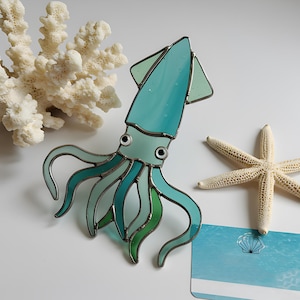 以下が含まれることがあります： ターコイズと緑色の色合いのステンドグラスのイカ。黒い目と銀色の輪郭があります。白いサンゴの枝とヒトデが白い表面のイカの隣にあります。貝殻のグラフィックが入った青いカードも存在します。
