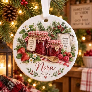 May include: A round white ceramic ornament with a white ribbon, featuring a printed illustration of two jars of red jam, a wooden spoon, strawberries, and pine branches. The text reads Good Things Grow Here, Canning Preserves Happier Days, Nora, and 2026.