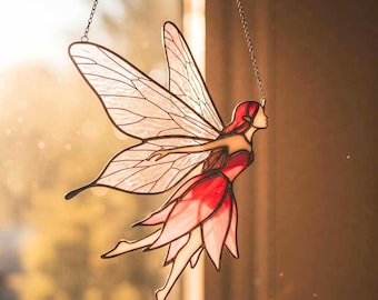 May include: A stained glass fairy, suspended by a chain, with red hair and a red and pink dress. The fairy has large, detailed wings and is posed in flight. The background is blurred, with sunlight filtering through.