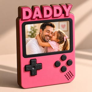 May include: A pink photo frame in the shape of a retro handheld game console, with the word "DADDY" in pink letters. The frame contains a photo of a man and a child. The console features black buttons and a directional pad.