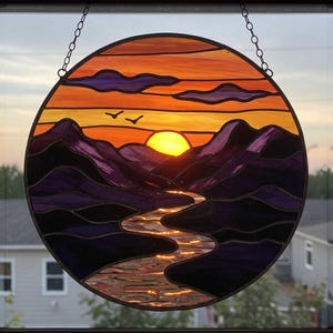 May include: A stained glass suncatcher depicting a sunset over purple mountains with a river winding through the valley. The suncatcher is round and has a wire hanger.