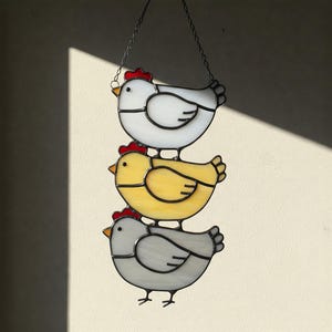 May include: A stained glass hanging decoration featuring three stylized chickens stacked vertically. The top chicken is white, the middle is yellow, and the bottom is gray. Each chicken has a red comb and black outlines. The piece hangs from a metal chain.