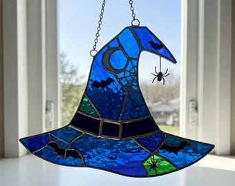 Puede incluir: Un sombrero de bruja de vitral colgado de una cadena. El sombrero es azul con una banda negra y presenta una luna creciente, telarañas y murciélagos. Hay dos arañas colgando del sombrero.