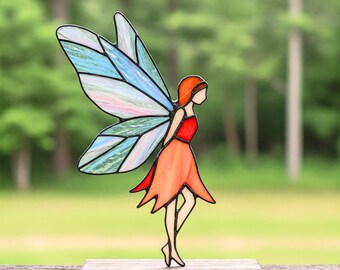 May include: A stained glass fairy figurine with blue and pink wings and an orange dress. The fairy is standing on a wooden base.