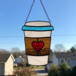 Könnte beinhalten: Glas-Suncatcher in Form einer Kaffeetasse mit rotem Herzdesign. Die Tasse hat einen weißen Deckel, einen blauen Rand und eine braune Manschette. Eine schwarze Kette ist zum Aufhängen befestigt. Für die Präsentation im Fenster konzipiert.