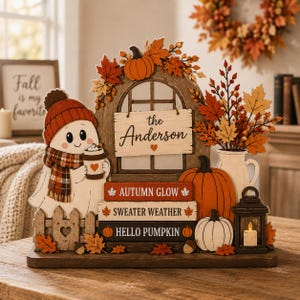 Può includere: Una decorazione da tavolo in legno con un fantasma che indossa un berretto rosso e una sciarpa a quadri mentre tiene una tazza di caffè. Include un cartello con scritto "the Anderson", libri con "AUTUMN GLOW", "SWEATER WEATHER", "HELLO PUMPKIN", zucche e una lanterna.