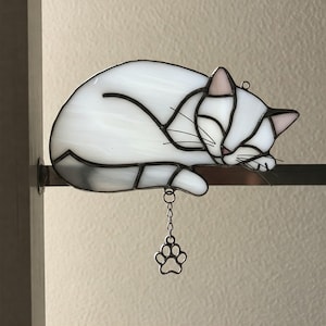 Puede incluir: Figura de gato de vidriera en blanco y gris, durmiendo plácidamente. El gato tiene orejas rosas y contornos negros. Una pequeña cadena con un dije de huella de pata cuelga debajo. La figura descansa sobre una barra de metal.