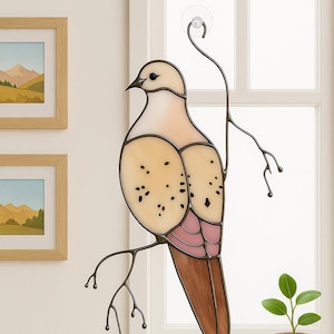 Op de afbeelding: Gebrandschilderde glazen duifdecoratie met een lichtbruin lichaam, roze accenten en zwarte details. De vogel zit op een metalen tak en is met een zuignap aan een raam bevestigd. Decoratieve kunst voor in huis.