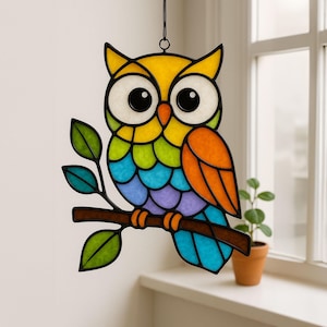 以下が含まれることがあります： カラフルなステンドグラスのフクロウの装飾。フクロウは、緑の葉が付いた茶色の枝にとまっています。フクロウの体は黄色、緑、青、オレンジ色です。フクロウは大きな丸い目を持ち、黒いワイヤーで吊り下げられています。