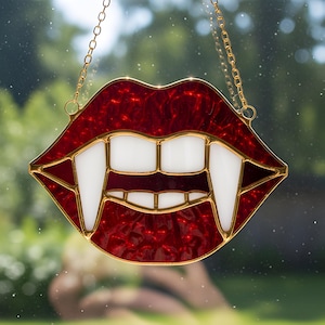 May include: A stained glass decoration in the shape of vampire lips. The lips are a deep red with white teeth and long fangs. The piece is outlined in gold and hangs from a gold chain. The background is a blurred view of greenery.
