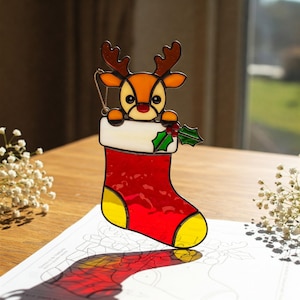 May include: A stained glass Christmas ornament featuring a reindeer peeking out of a red stocking with yellow accents. The reindeer has brown antlers and a red nose. A sprig of holly is on the stocking. The ornament hangs from a gold chain.