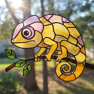 May include: A stained glass chameleon suncatcher in yellow, pink, and purple, perched on a brown branch with green leaves. The chameleon has a detailed, segmented design and a curled tail. Sunlight shines through the glass, creating a colourful display.