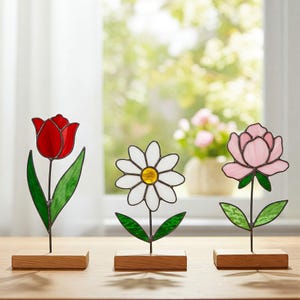 May include: Three stained glass flower sculptures: a red tulip, a white daisy with a yellow center, and a pink peony. Each flower is mounted on a black metal stem and set in a small, rectangular wooden base. The background is a blurred view of a window.