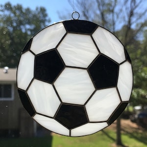 Pode incluir: Uma bola de futebol em vitral com pentágonos e hexágonos pretos e brancos. O vidro é emoldurado com uma borda escura e tem um pequeno laço de metal para pendurar. A bola de futebol é um item decorativo.