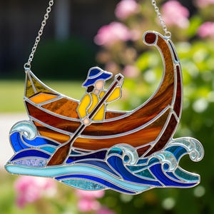 May include: Stained glass suncatcher depicting a person rowing a boat. The boat is brown and orange, with a figure in yellow and blue. The waves are blue and turquoise. The suncatcher hangs from a silver chain.