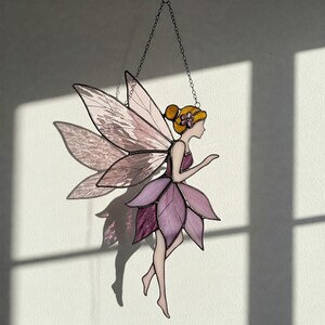 May include: A stained glass fairy with pink wings and a purple dress. The fairy is hanging from a chain and has a yellow flower in her hair.