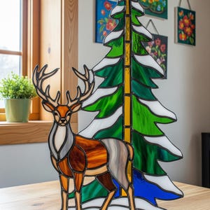 May include: Stained glass artwork depicting a deer and a Christmas tree. The deer is coloured in shades of brown, orange, and grey, and the tree is green with white detailing. The piece is mounted on a wooden base, suitable for display as a decorative item.