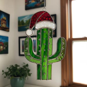 May include: A stained glass cactus ornament with a red Santa hat and white trim. The cactus is green with silver accents. The ornament is hanging in front of a window.