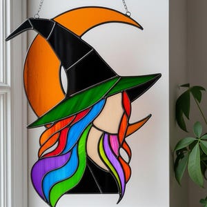 Könnte beinhalten: Buntglas-Hexe mit schwarzem Hut, grünem Rand und bunten Haaren vor einem orangefarbenen Halbmond. Das Haar der Hexe ist regenbogenfarben. Ein dekoratives Stück für Halloween.