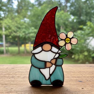 Peut inclure: Figurine de gnome en vitrail avec un chapeau rouge, tenant une fleur. Le gnome a une barbe blanche, un nez bronzé et une tenue bleu-vert. La fleur a des pétales roses et jaunes.