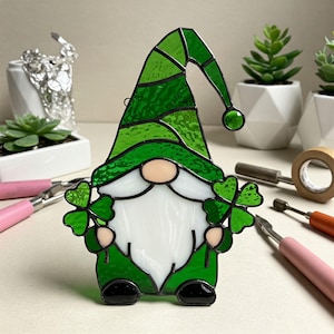 May include: A stained glass gnome with a green hat and beard, holding shamrocks in each hand. The gnome is green and black with a white beard.