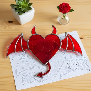 May include: A red stained glass heart with devil horns, wings, and a tail. The heart is on top of a drawing of hearts with wings. A small succulent in a white pot and a red rose in a glass vase are in the background.
