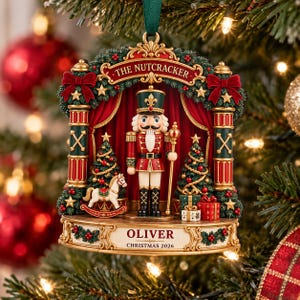 May include: A decorative Christmas ornament featuring a nutcracker soldier standing on a stage with red curtains, a rocking horse, and gifts. The text reads THE NUTCRACKER, OLIVER, and CHRISTMAS 2026. It is framed by green garlands and gold pillars.