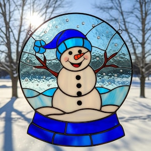 May include: Stained glass snowman in a snow globe design. The snowman has a blue hat, carrot nose, and twig arms. The snow globe is round with a blue base and a winter scene inside. The piece is designed to hang.