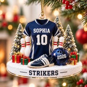 May include: A bowling-themed Christmas ornament featuring a navy blue jersey with the name SOPHIA and number 10, a bowling ball marked 2026, bowling pins, a bowling shoe, and gift boxes on a snowy base with the text STRIKERS printed on the front.