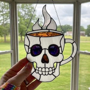 Puede incluir: Un vitral colgante presenta una calavera blanca con forma de taza de café con una llama en la parte superior. La taza tiene un líquido naranja en su interior y ojos morados.