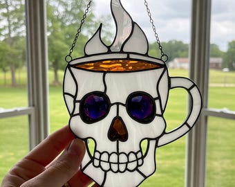 Puede incluir: Un vitral colgante presenta una calavera blanca con forma de taza de café con una llama en la parte superior. La taza tiene un líquido naranja en su interior y ojos morados.