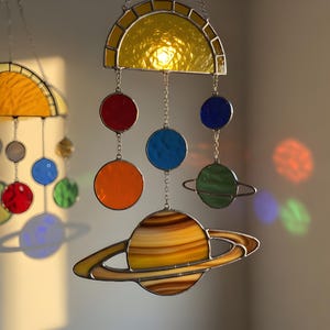 Puede incluir: Móvil del sistema solar de vidrieras con un sol amarillo, planetas en rojo, naranja, azul y verde, y un planeta con anillos. Los planetas están suspendidos por cadenas. El sol tiene forma de semicírculo.