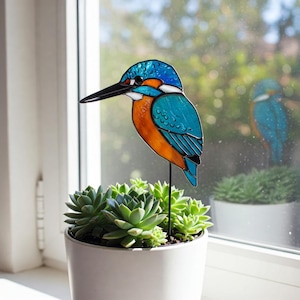 Puede incluir: Una figurita de pájaro martín pescador de vidrio coloreado con colores azul, naranja y verde. La figurita está posada en una estaca de metal negro y se coloca en una maceta blanca con una planta suculenta.