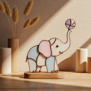 Peut inclure: Figurine d'éléphant en vitrail aux couleurs pastel. L'éléphant tient une balle assortie. La figurine est montée sur un socle en bois, créant un élément décoratif charmant et coloré.