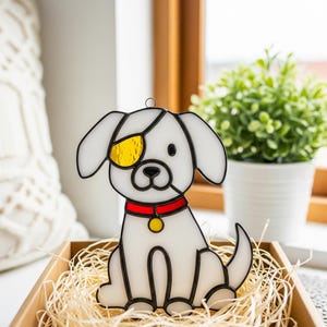 May include: A white stained glass dog with a black outline, wearing a yellow eyepatch and a red collar with a gold pendant. The dog is sitting in a wooden box filled with straw. A decorative item.