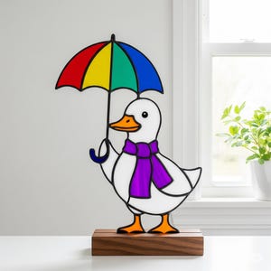 May include: A whimsical stained glass duck figurine holding a rainbow umbrella. The duck wears a purple scarf and has orange feet and beak. It stands on a wooden base, perfect for home decor.