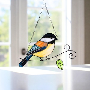 May include: Stained glass bird suncatcher in black, white, orange, blue, and yellow. The bird sits on a metal branch with a green leaf. It hangs from a chain, suitable for window or wall decoration. A decorative piece.