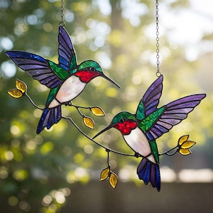 Könnte beinhalten: Zwei Buntglas-Kolibries mit schillernden Flügeln und roten, grünen und blauen Akzenten sitzen auf einem Ast mit gelben Blättern. Die Kolibris hängen an einer Kette.