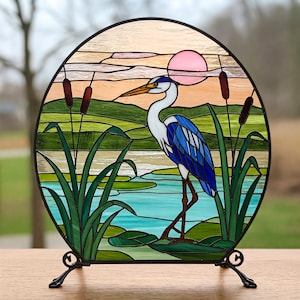 May include: A stained glass window depicting a blue heron standing in a marsh with cattails and a pink sunset in the background.