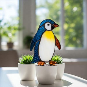 Può includere: Una statuina di pinguino in vetro colorato con testa blu, petto giallo e pancia bianca. Il pinguino ha zampe arancioni e un becco nero. È esposto su tre piccoli vasi bianchi con succulente verdi.