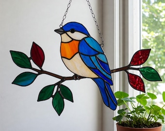 May include: A stained glass bluebird ornament, featuring a blue, orange, and white bird on a brown branch with green and red leaves. It hangs from a black chain. A decorative piece for a window, measuring approximately 20 cm in length.