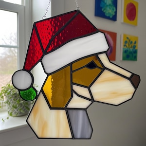 May include: Stained glass dog head wearing a Santa hat. The dog's head is made of various colors of glass, including red, white, yellow, brown, and gray. The hat is red with a white brim and a white pom-pom. The piece is hanging from a chain.
