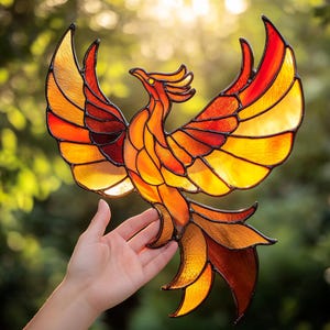 Peut inclure: Un phénix en vitrail avec des pièces de verre orange, jaune et rouge. Le phénix est en vol, les ailes déployées. Le vitrail est placé sur un fond flou de feuillage vert.