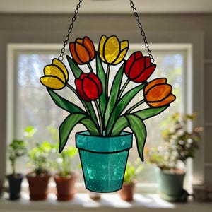 May include: Stained glass tulip flower arrangement in a teal pot, hanging from a chain. The tulips are red, orange, and yellow with green leaves. The window behind the flowers is in soft focus.