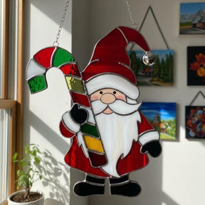 Peut inclure: Décoration de Noël en vitrail représentant le Père Noël tenant une canne en bonbon. Le personnage est principalement rouge avec des bordures blanches, une barbe blanche et un visage rose. La canne en bonbon est rouge, verte, jaune et blanche.