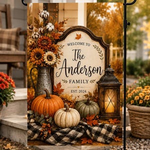 May include: A rectangular garden flag featuring a fall-themed display with a metal pitcher of sunflowers and cotton, pumpkins, and a lantern. The center sign reads: Welcome to The Anderson Family EST. 2024. A black and white buffalo plaid cloth sits below.