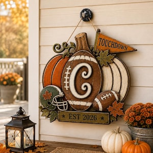 May include: A decorative wooden wall hanging shaped like a pumpkin and football, featuring the letter C, a green football helmet, a pennant with the text TOUCHDOWN, and a rectangular plaque at the bottom with the text EST 2026 in brown and orange.